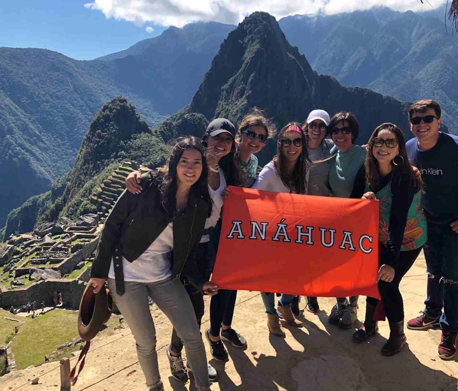 Grupo de personas sonriendo y sosteniendo una bandera naranja con la palabra “Anáhuac” frente a las ruinas de Machu Picchu y montañas verdes.